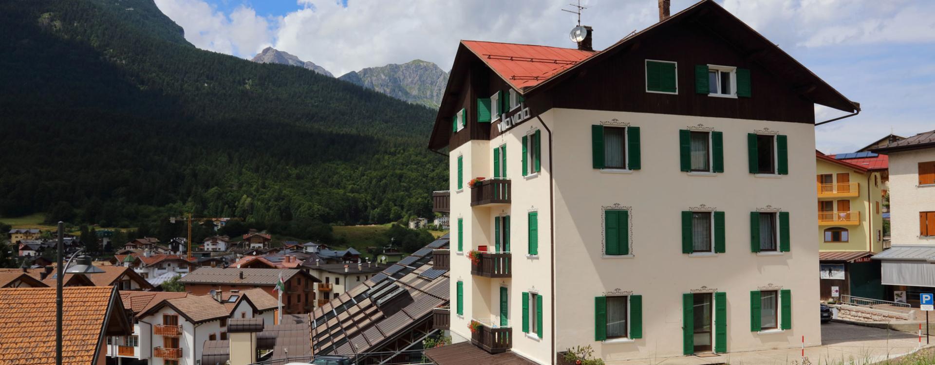 Edificio con persiane verdi in un villaggio di montagna, circondato da colline e case.