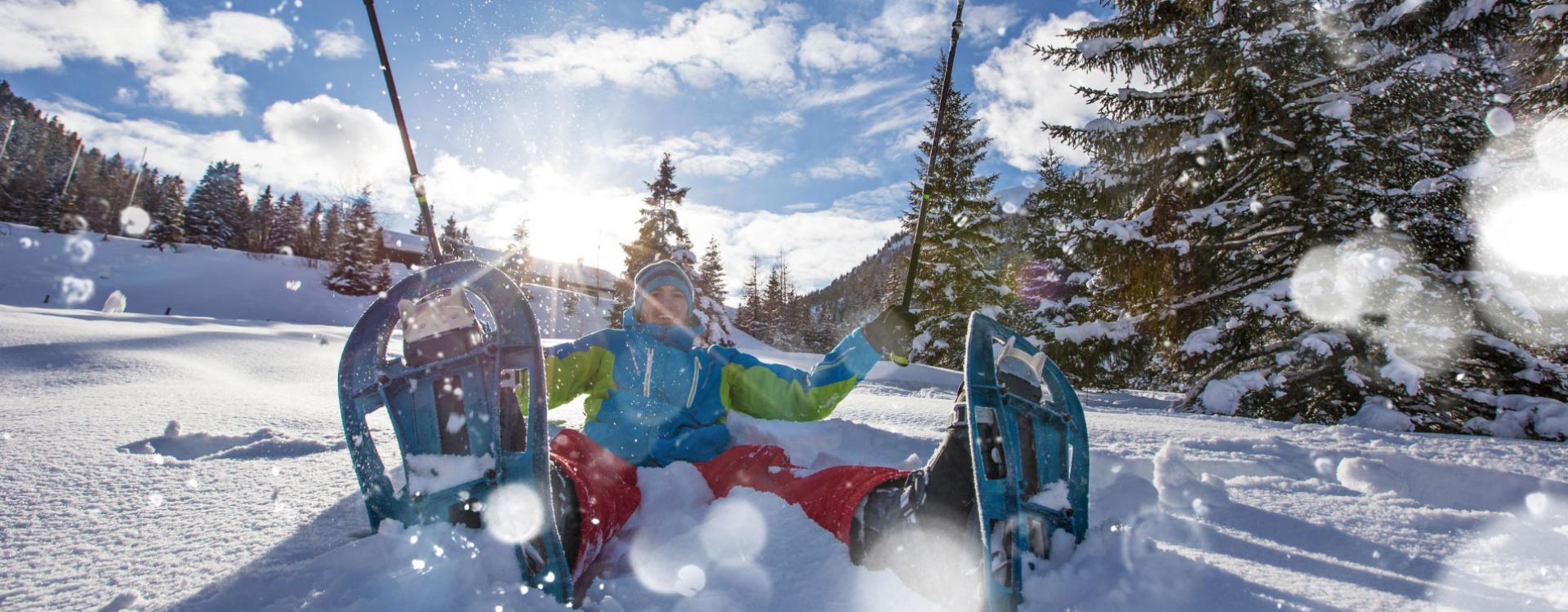 Person with snowshoes, sitting in the snow, sun and trees in the background.