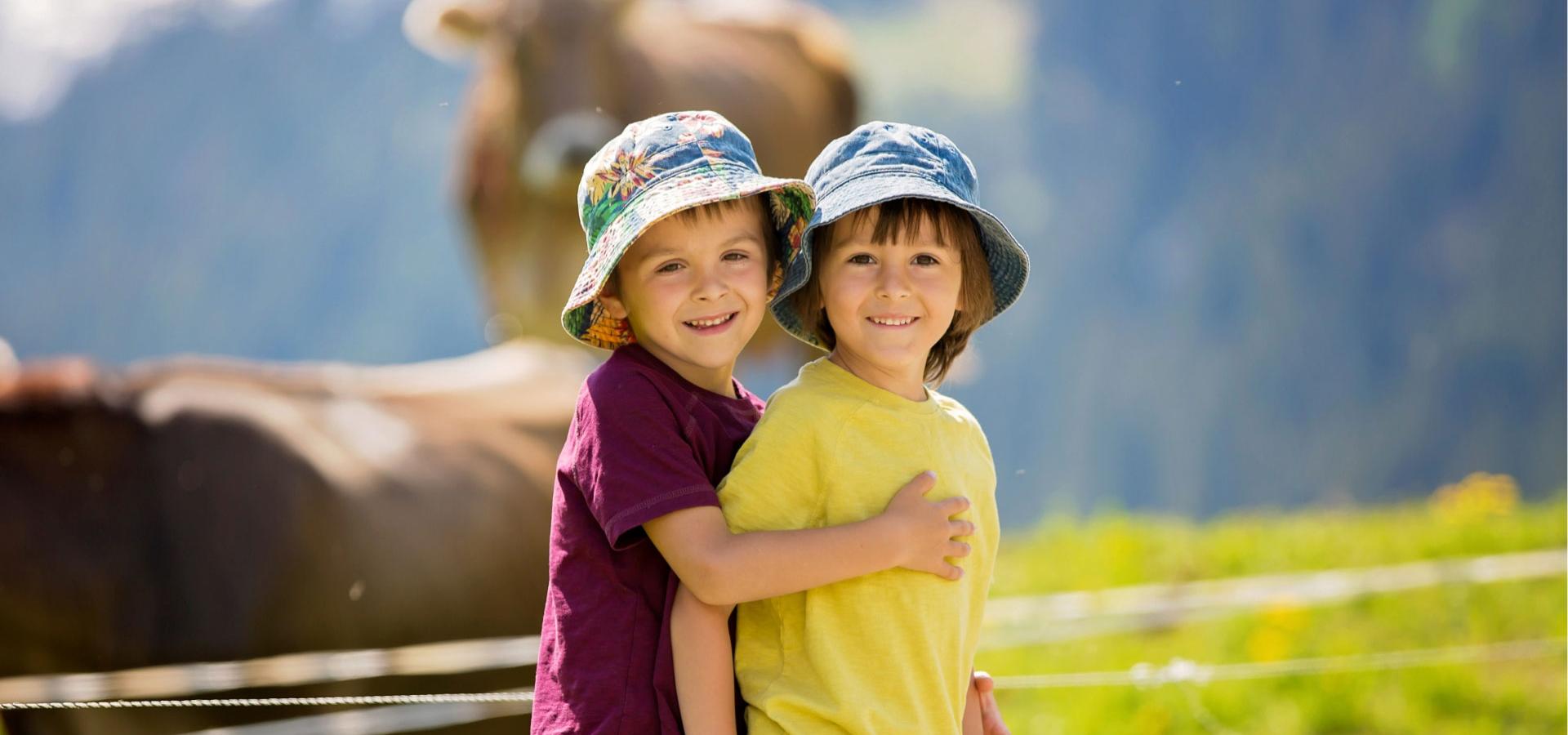 Two smiling children with colorful hats in a meadow with a cow in the background.