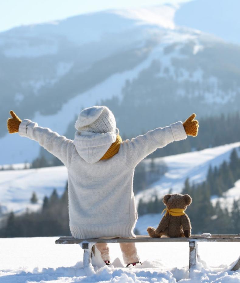 Child in snow with teddy bear, arms open towards snowy mountains.